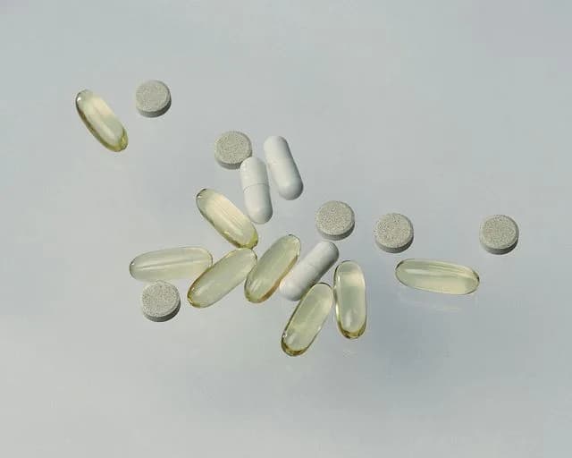 Pale amber softgel capsules, white capsules and small round tablets scattered across a pale grey surface