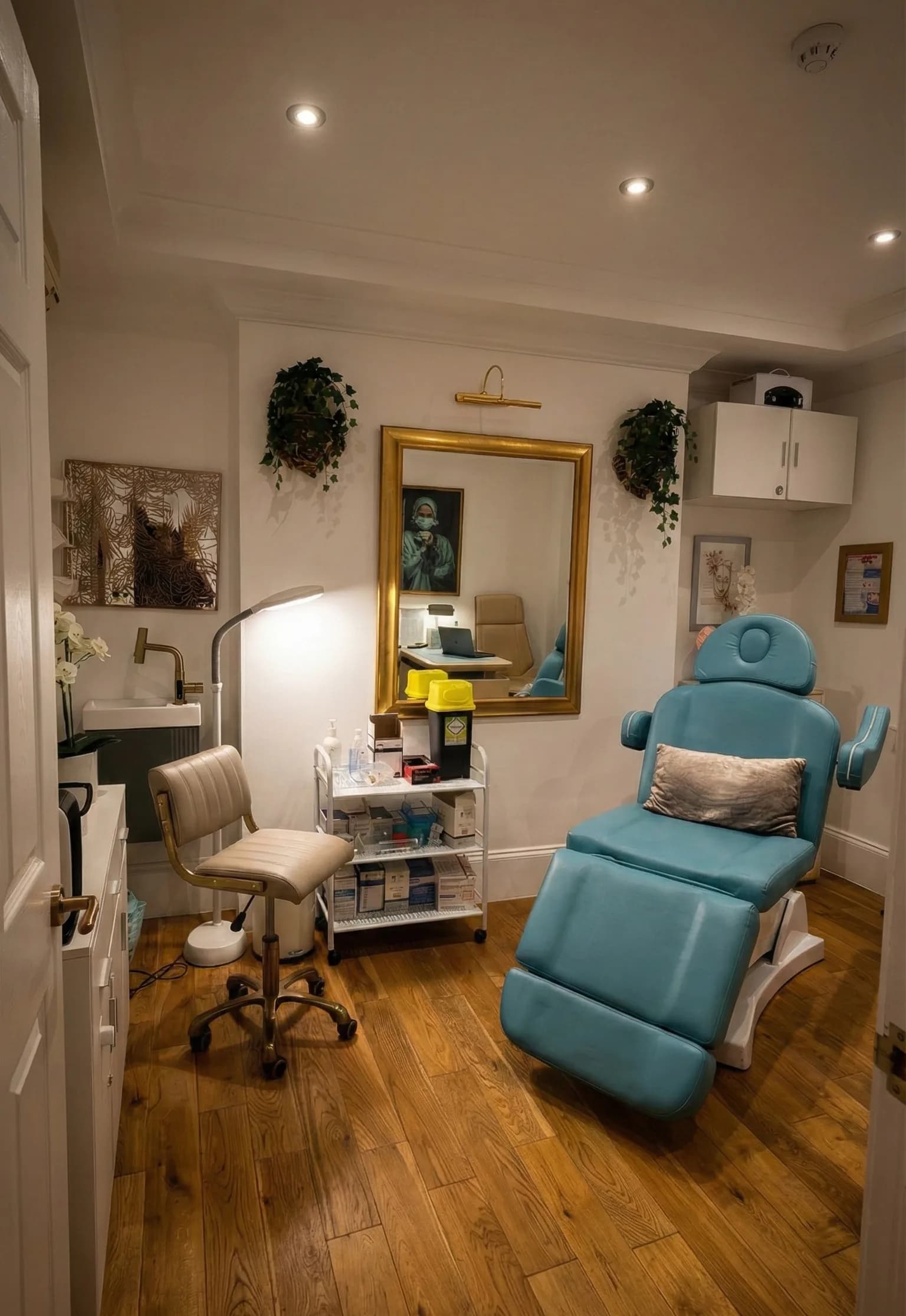 A treatment room at the LV8 Health London clinic: a reclining treatment chair, an instrument trolley and a gold framed mirror on a white wall